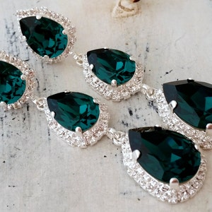May include: A pair of long, dangling earrings with three teardrop-shaped emerald green crystals. Each crystal is surrounded by a halo of smaller, clear crystals.