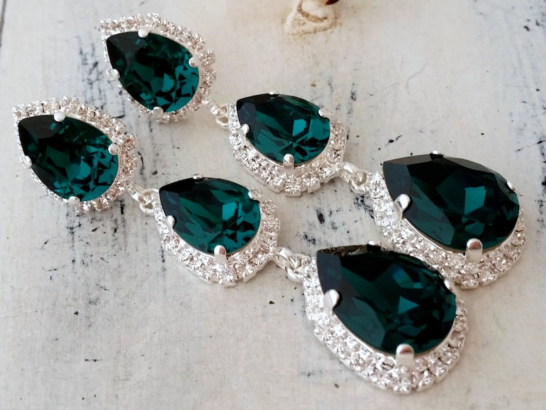 May include: A pair of long, dangling earrings with three teardrop-shaped emerald green crystals. Each crystal is surrounded by a halo of smaller, clear crystals.