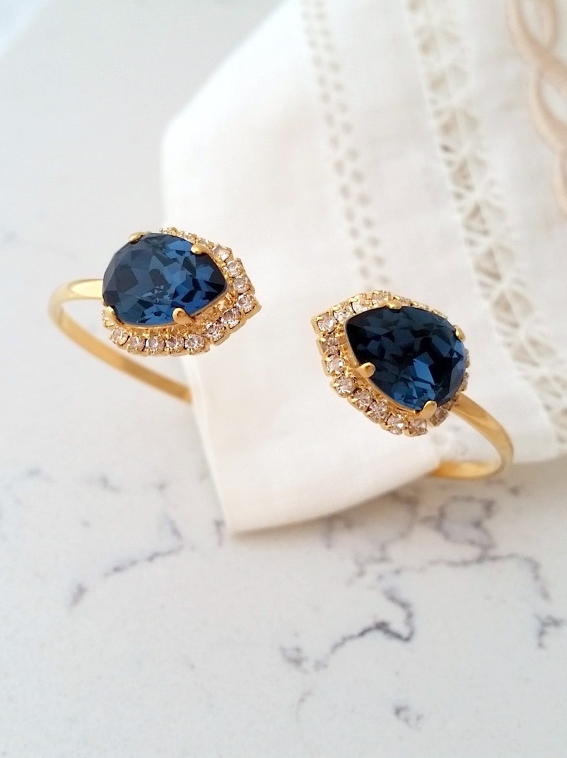 May include: A gold bangle bracelet with two teardrop-shaped blue gemstones surrounded by smaller clear rhinestones.