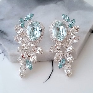 May include: A pair of silver earrings with a large, oval-shaped light blue crystal in the center of each earring. The crystals are surrounded by smaller, clear crystals that are arranged in a floral pattern. The earrings are dangling and have a long, teardrop shape.