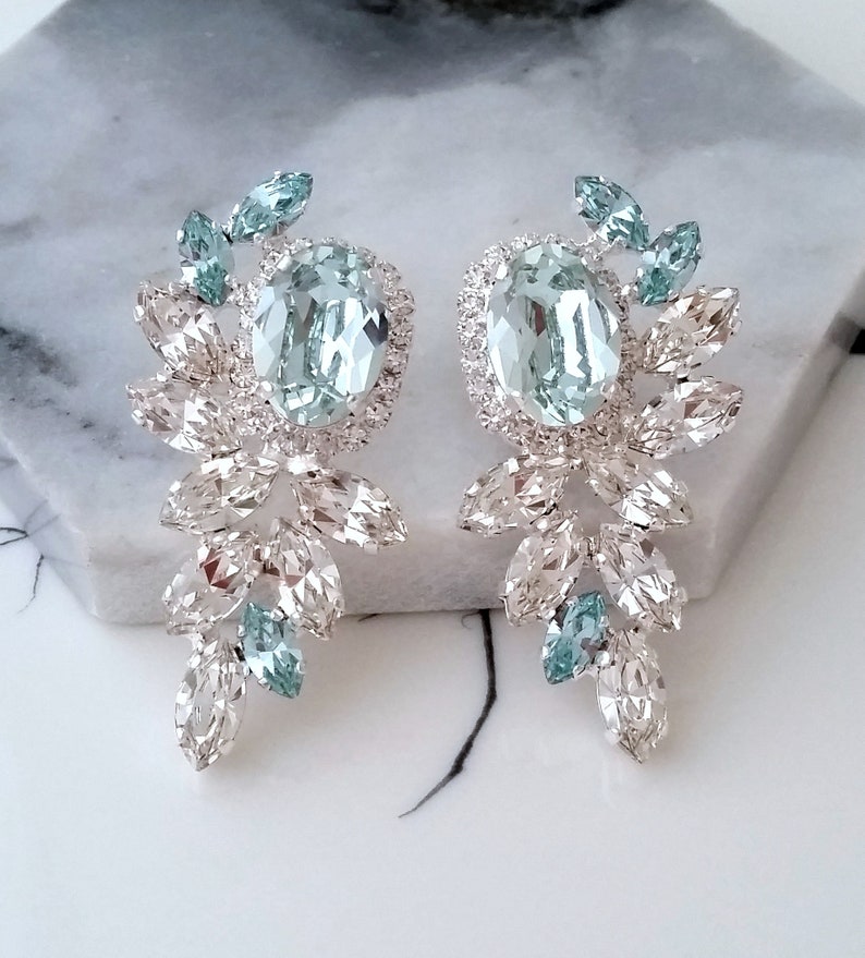 May include: A pair of silver earrings with a large, oval-shaped light blue crystal in the center of each earring. The crystals are surrounded by smaller, clear crystals that are arranged in a floral pattern. The earrings are dangling and have a long, teardrop shape.