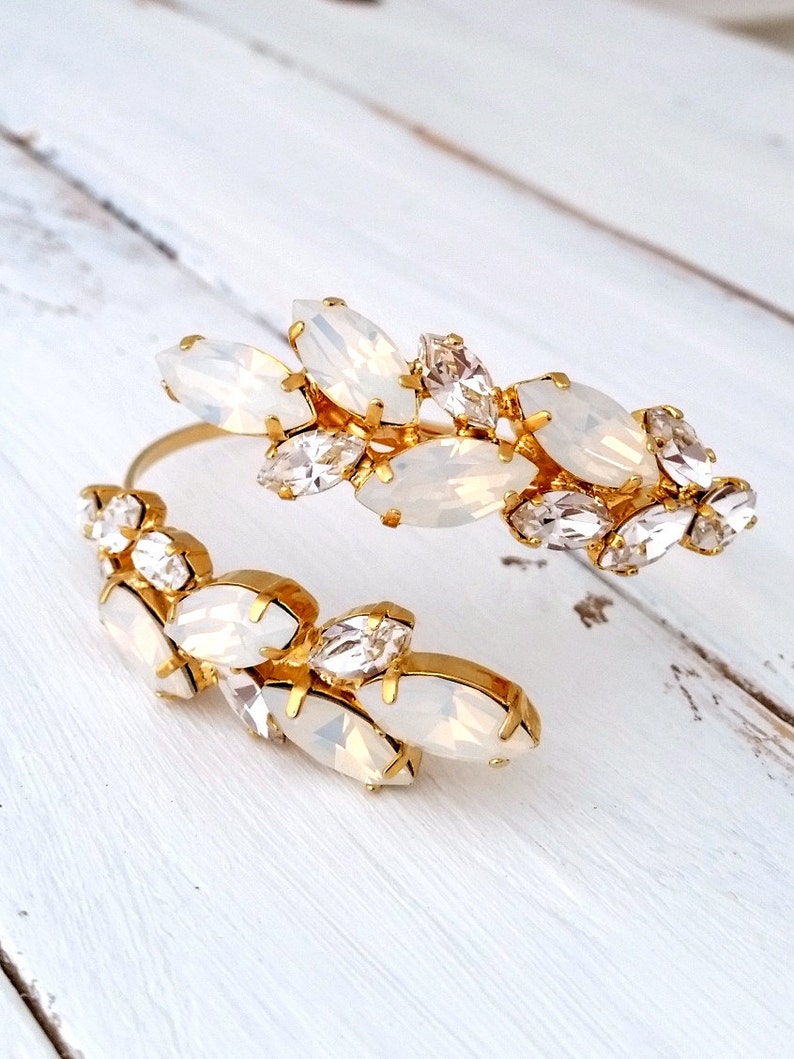 May include: A gold cuff bracelet with a cluster of white and clear crystal stones. The stones are arranged in a floral pattern.