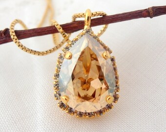 May include: A gold-toned necklace featuring a large, pear-shaped pendant. The pendant has a light amber-colored center stone, surrounded by a border of smaller, clear crystals. The necklace chain is also gold-toned, and the pendant is suspended from a small loop.
