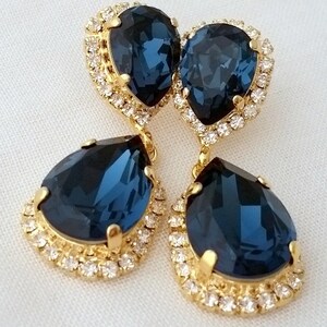May include: A pair of dangle earrings with a teardrop-shaped blue crystal surrounded by a halo of smaller clear crystals. The earrings are gold-toned.