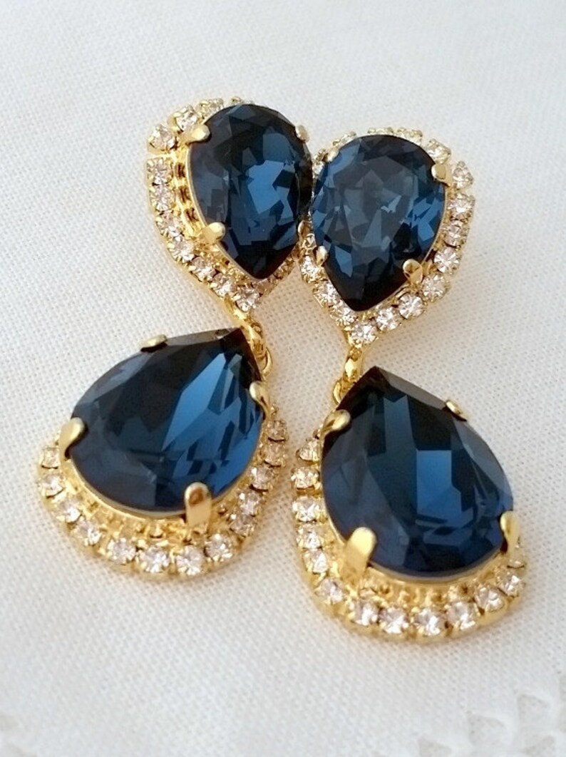 May include: A pair of dangle earrings with a teardrop-shaped blue crystal surrounded by a halo of smaller clear crystals. The earrings are gold-toned.