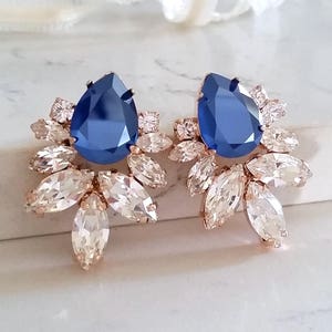 May include: A pair of rose gold earrings with a large blue teardrop-shaped crystal surrounded by smaller clear crystals.