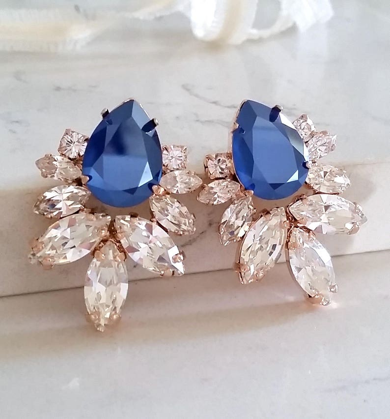May include: A pair of rose gold earrings with a large blue teardrop-shaped crystal surrounded by smaller clear crystals.
