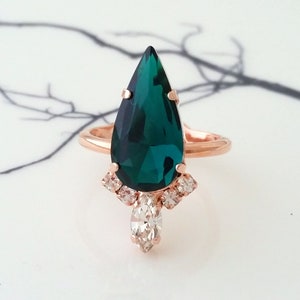 May include: A pear-shaped emerald green gemstone ring with a rose gold band. The ring has a cluster of smaller clear gemstones around the main stone and a single pear-shaped clear gemstone hanging below.