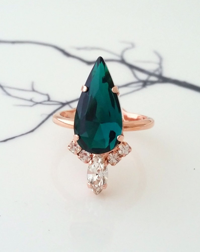 May include: A pear-shaped emerald green gemstone ring with a rose gold band. The ring has a cluster of smaller clear gemstones around the main stone and a single pear-shaped clear gemstone hanging below.