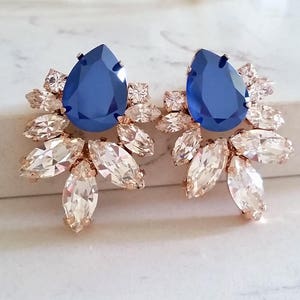 May include: A pair of rose gold earrings with a large blue teardrop-shaped crystal in the center of each earring. The crystals are surrounded by smaller clear crystals in a floral design.