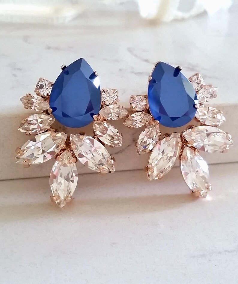 May include: A pair of rose gold earrings with a large blue teardrop-shaped crystal in the center of each earring. The crystals are surrounded by smaller clear crystals in a floral design.