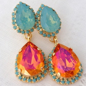 May include: A pair of dangle earrings with two teardrop-shaped crystals. The top crystal is light blue and the bottom crystal is orange with pink and gold iridescent accents. Both crystals are surrounded by a gold-toned setting with a turquoise blue border.