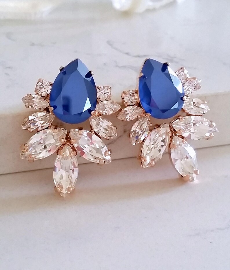 May include: A pair of rose gold earrings with a large blue teardrop-shaped crystal surrounded by smaller clear crystals.
