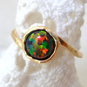 May include: A gold ring with a round, multi-colored opal gemstone set in a bezel setting. The ring is on a white crocheted surface.