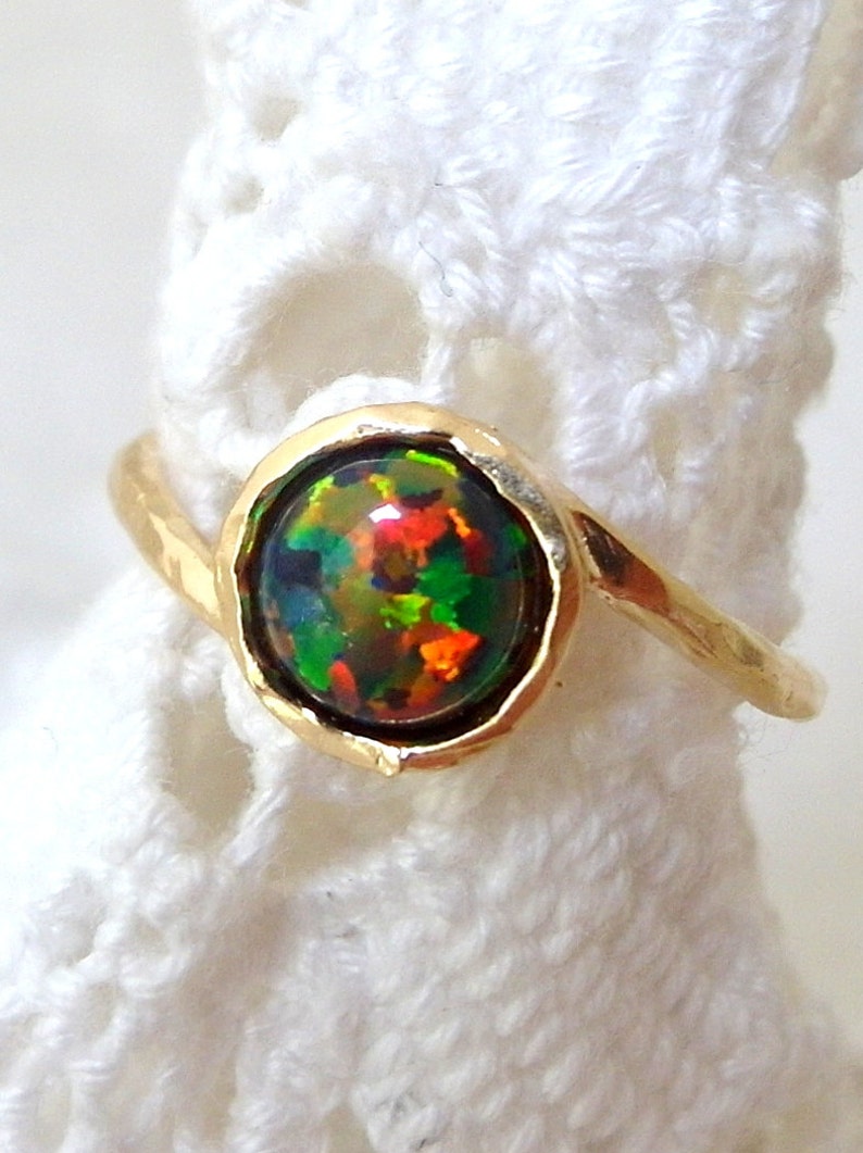 May include: A gold ring with a round, multi-colored opal gemstone set in a bezel setting. The ring is on a white crocheted surface.
