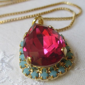 May include: A gold chain necklace with a teardrop-shaped pendant. The pendant features a large, bright pink crystal surrounded by a halo of smaller, turquoise crystals.