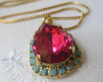 May include: A gold chain necklace with a teardrop-shaped pendant. The pendant features a large, bright pink crystal surrounded by a halo of smaller, turquoise crystals.