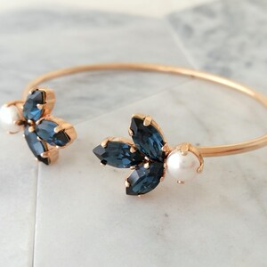 May include: A gold bangle bracelet with two blue crystal flower accents and a pearl on each side.