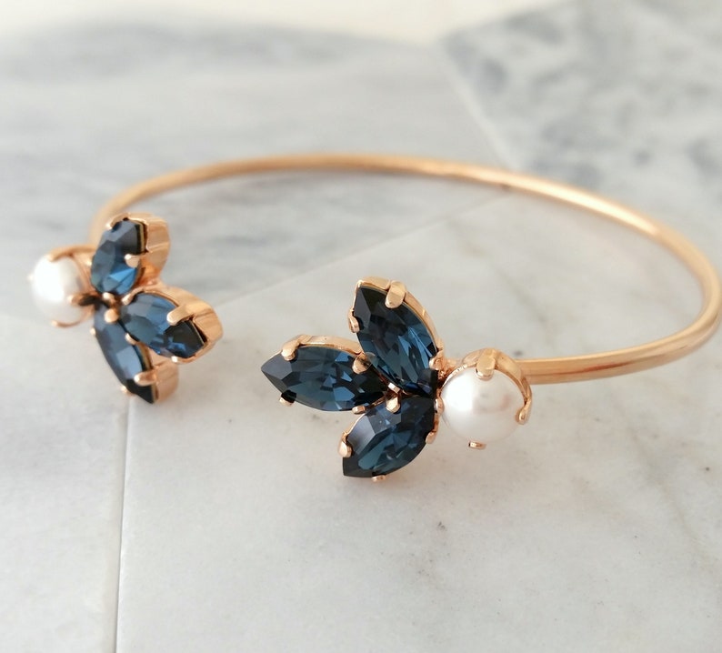 May include: A gold bangle bracelet with two blue crystal flower accents and a pearl on each side.