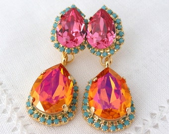 May include: A pair of dangle earrings with two teardrop-shaped crystals. The top crystal is pink and the bottom crystal is orange with a pink iridescent sheen. The crystals are surrounded by a gold-colored metal setting with a turquoise-colored trim.