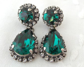 May include: A pair of dangle earrings with emerald green pear-shaped stones surrounded by a halo of smaller clear stones.