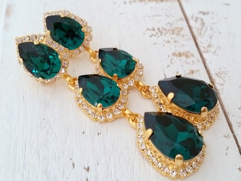 May include: Emerald green teardrop earrings with gold settings and clear crystal accents. The earrings are long and dangle.