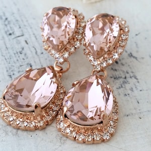 May include: Rose gold dangle earrings with two teardrop shaped blush pink crystals. The crystals are surrounded by a halo of smaller clear crystals.