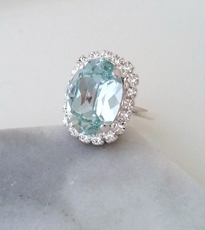 May include: A silver ring with a large oval-shaped light blue gemstone surrounded by a halo of smaller clear rhinestones.
