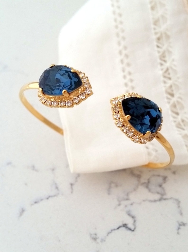 May include: Gold bangle bracelet with two teardrop-shaped blue gemstones surrounded by clear rhinestones.