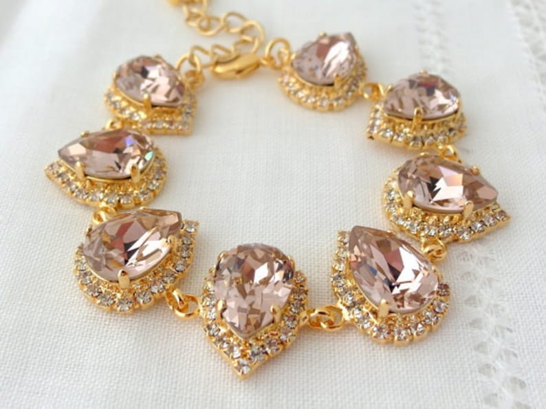 May include: A gold bracelet with a teardrop-shaped crystal design. The crystals are a light blush pink color and are set in a gold metal frame. The bracelet is made of a chain link design.