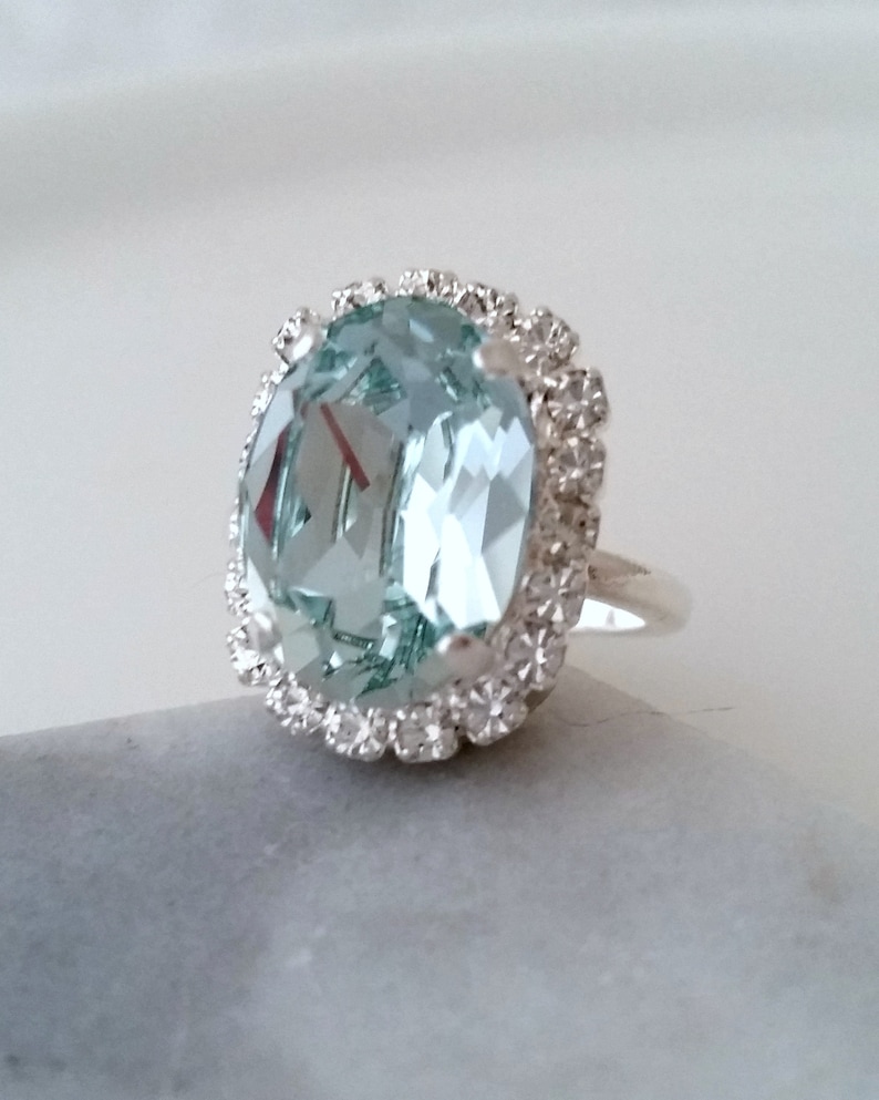May include: A silver ring with a large, oval-shaped, light blue gemstone surrounded by a halo of smaller, clear gemstones.