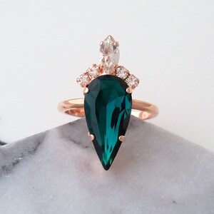 May include: A rose gold ring with a large pear-shaped emerald green gemstone and smaller clear gemstones around the band.