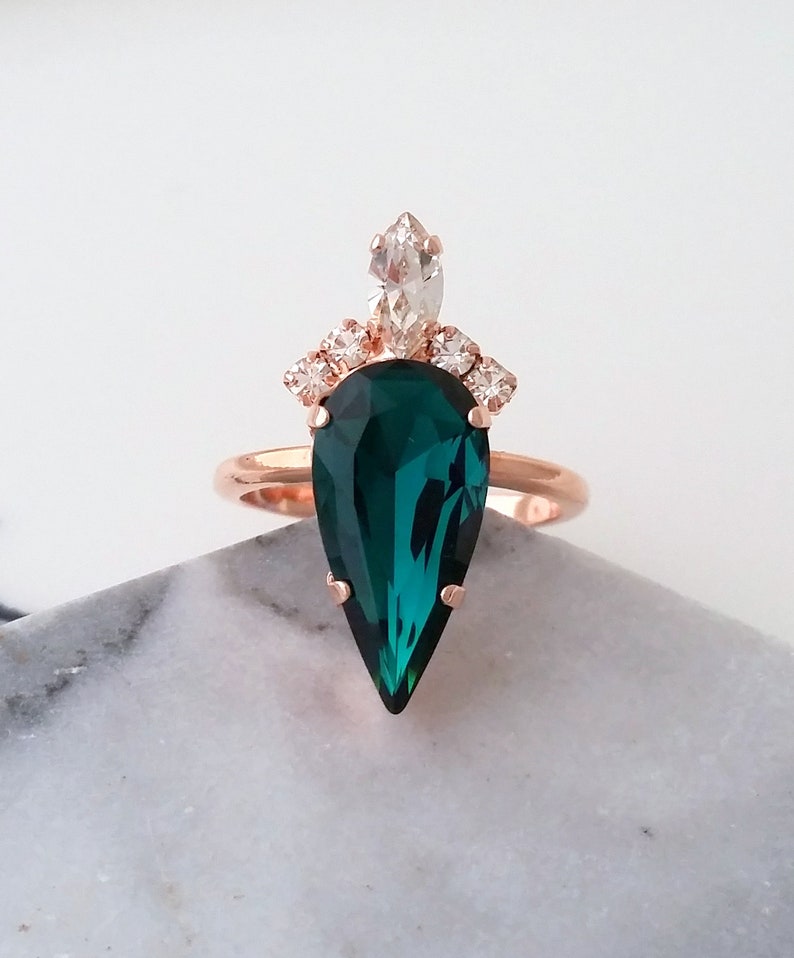 May include: A rose gold ring with a large pear-shaped emerald green gemstone and smaller clear gemstones around the band.