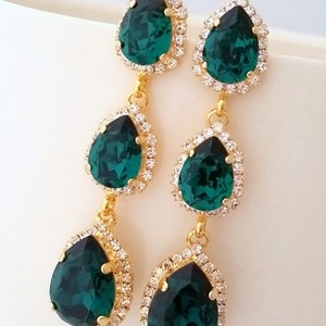 May include: Emerald green teardrop earrings with gold settings and a halo of clear rhinestones. The earrings are three-tiered with a smaller teardrop at the bottom.