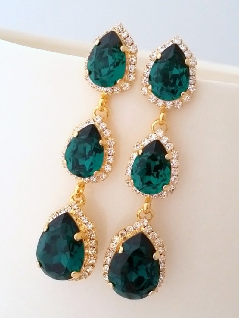 May include: Emerald green teardrop earrings with gold settings and a halo of clear rhinestones. The earrings are three-tiered with a smaller teardrop at the bottom.