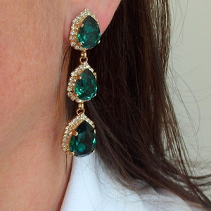 May include: A pair of gold dangle earrings with three emerald green teardrop-shaped stones. Each stone is surrounded by a halo of smaller clear stones.