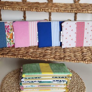 Peut inclure: Une collection de pochettes zippées rectangulaires en tissu avec divers motifs, notamment des imprimés floraux, rayés, cerises et botaniques dans des tons rose, bleu, vert, jaune et rouge. Chaque pochette porte une étiquette ATELIER EPHAE.