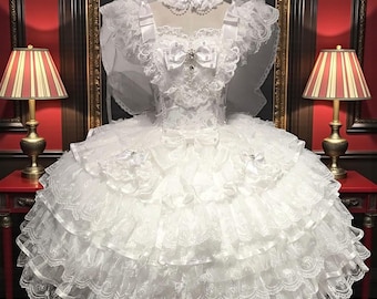 Op de afbeelding: Een witte trouwjurk met een gelaagde rok en ingewikkelde kanten details. Het lijfje heeft een strik en versieringen. Een sluier en kraag maken het ensemble compleet. De jurk wordt getoond op een mannequin, geflankeerd door lampen.