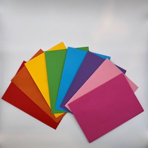Pode incluir: Um leque de folhas de papel retangulares coloridas, que vão do vermelho ao laranja, amarelo, verde, azul, roxo e rosa. As folhas estão dispostas em semicírculo, mostrando um espectro de cores vibrantes.