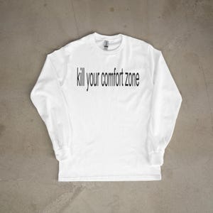Könnte beinhalten: Weißes Langarmshirt mit dem schwarzen Schriftzug "kill your comfort zone". Das Kleidungsstück liegt flach auf einer grauen Oberfläche. Das Shirt hat einen Rundhalsausschnitt und lange Ärmel.