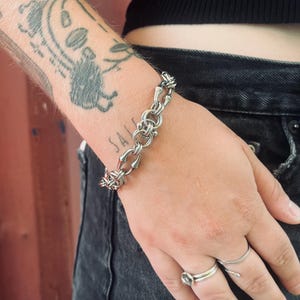 May include: A silver-toned chain bracelet featuring a series of interlocking circular and elongated links. The bracelet has a metallic finish and a secure clasp mechanism, worn around the wrist. It is accompanied by two silver-toned rings on the fingers.