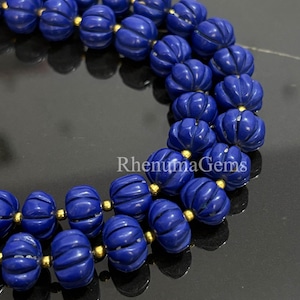Peut inclure: Gros plan sur un collier avec des perles sculptées bleu foncé en forme de citrouille. Les perles sont enfilées avec des entretoises dorées. Le collier est présenté sur une surface noire réfléchissante. Le texte "Rhenuma Gems" est visible.