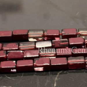 Könnte beinhalten: Ein Strang quadratischer Perlen in einem tiefen Kastanienrot mit metallisch reflektierender Oberfläche. Die Perlen sind in horizontalen Reihen angeordnet und weisen eine glatte, polierte Oberfläche mit einigen sichtbaren Lichtreflexen auf.