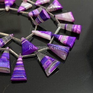 May include: Close-up of a strand of purple and white gemstone beads. The beads are triangular and feature a striped pattern. The text "RhenumaGems" is visible on the image. These beads are suitable for jewelry making.
