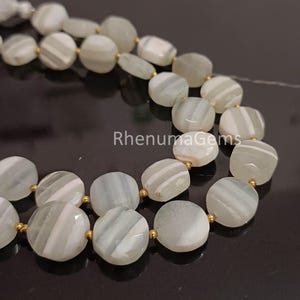May include: Strands of light gray and white striped gemstone beads with gold-colored spacers. The beads are round and faceted, with a polished finish. The text "RhenumaGems" is visible.