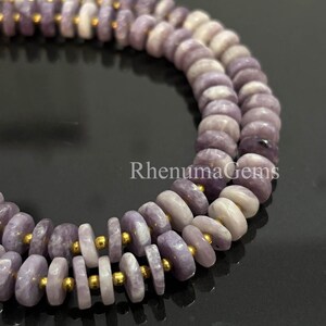 Puede incluir: Primer plano de dos hilos de cuentas de piedras preciosas de color lavanda. Las cuentas en forma de disco están separadas por pequeños espaciadores dorados. Las cuentas tienen un acabado pulido y están ensartadas, creando un collar o pulsera.