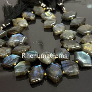 Könnte beinhalten: Strang aus facettierten Labradorit-Edelsteinperlen. Die Perlen sind eine Mischung aus Grau-, Blau- und Goldtönen mit einer geometrischen Form. Jede Perle wird durch eine kleine goldfarbene Perle getrennt. Schwarze Quasten sind oben zu sehen.