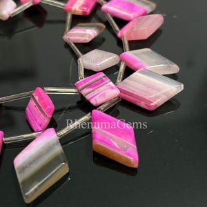 May include: Close-up of a strand of pink and gray agate gemstone beads. The beads are diamond-shaped and strung on a silver wire. The beads have a polished finish and are arranged against a dark background. The text "RhenumaGems" is visible.