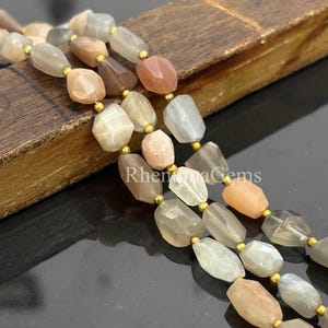 Op de afbeelding: Strengen gefacetteerde edelsteen kralen in tinten perzik, grijs en crème, met goudkleurige accenten. De kralen zijn aan elkaar geregen, waardoor een ketting of armband ontstaat. De edelstenen hebben een gepolijste, onregelmatige vorm.