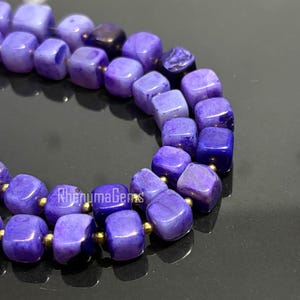 May include: Close-up of a necklace made of square, polished, purple beads. The beads are strung together with gold-colored spacers. The necklace is displayed on a dark, reflective surface. The text "RhenumaGems" is visible.